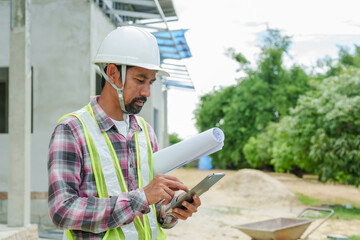 Wall Mural - Asian Construction worker in plaid shirt and reflective vest holds blueprints and tablet, standing at a building site with greenery around. Architects and engineers supervise house construction.