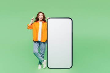 Wall Mural - Full body young woman wear orange shirt white t-shirt casual clothes big huge blank screen mobile cell phone smartphone with area point finger up isolated on plain green background. Lifestyle concept.