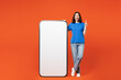 © ViDi Studio - Full body young woman wear blue t-shirt casual clothes big huge blank screen mobile cell phone smartphone with workspace copy space area point index finger up isolated on plain red orange background.