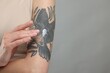 Tattooed woman applying cream onto her arm on gray background, closeup. Space for text