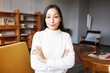 Indoor portrait of charming confident asian woman psychologist in beige sweater smiling at camera sitting with crossed hands on sofa in cozy cabinet, satisfied with her work and results