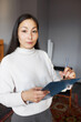 Indoor portrait of pretty female designer standing with clipboard in hands, examining living-room to make new project, making notes, looking at camera with confident facial expression