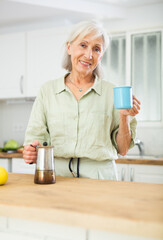 Wall Mural - Positive old woman drinking coffee in kitchen at home. Senior woman with mug of hot drink.