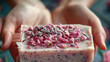 A close-up view capturing a person holding a handmade soap bar topped with vibrant dried flowers against a blurred background.