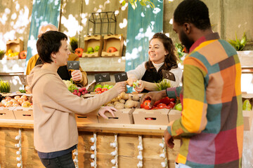 Wall Mural - Smiling friendly merchant offering customer to taste varieties of fresh seasonal apples at food market. Happy female farmer selling local homegrown organic fruits and vegetables.