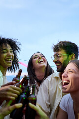 Wall Mural - Vertical. Cheerful multiracial young people toast with beer bottles gathered celebrating laughing on summer outdoors. Excited group happy millennial friends together enjoying drinks at rooftop party