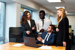 © artiemedvedev - Multiethnic business team engages in lively discussion around laptop in modern office setting, with smiling woman standing, African man with papers, businessman gesturing, blonde woman listening.
