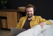 A man wearing headphones and a yellow jacket is sitting on a couch with a laptop in front of him. He is smiling and he is enjoying his time
