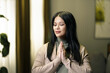 A woman is praying in a room with a plant in the background. She is wearing a brown sweater and has her hands clasped together