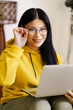 A woman wearing glasses and a yellow hoodie is sitting on a couch and looking at a laptop