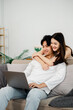 © Satori Studio - Asian couple shows closeness and comfort, embracing each other while focused on a laptop on their cozy sofa at home.