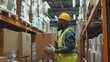 © Kartika - Afro male wearing safety outfit inspects cardboard box for shipment. Warehouse worker checking stocks in large warehouse.