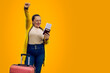 Beautiful woman in summer clothes with pink suitcase, happy with her hand up, celebrating her cheap ticket on yellow background. Tourist very happy to travel abroad with a bargain ticket. Copy space.