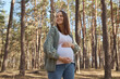 Attractive smiling brown haired Caucasian pregnant woman relaxing in spring forest expectant mother with stroking her belly enjoying beautiful nature while walking in woodland