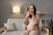 Sick ill Caucasian pregnant woman sitting on sofa at home having sore throat using spray for medical treatment taking care of her health during pregnancy