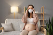 Sick pregnant woman wearing medical mask suffering terrible headache and flu symptoms massaging her temples while sitting on couch in cozy living room at home