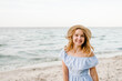 Happy beautiful girl standing on beach ocean and enjoying sunny summer day on vacation. Young smiling blond woman in straw hat walks near sea and looks at camera. Portrait of stylish female on sand.