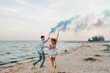 Man and woman having fun running on sand and holding hands blue and pink color smoke bombs and looking at each other on beach. Happy married couple. Baby shower party. Twins boy and girl. Back view