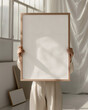 © Amy - A woman female artist holding a blank wood oak thin framed 20x28, 50x70 art print mockup, in a minimal neutral interior, with dappled sunlight from window