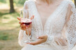 The bride is holding a glass of red wine outdoors. The girl has a beautiful dress and neckline closeup. Wedding ceremony and backyard party. Tasting wine. Female drinking wine closeup.