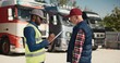Side view of two multiethnic workers speaking with each other about load and unload freight at warehouse. Actively speaking while standing at lorries parking in sunny day. Shipping concept.