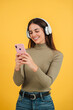 © Jose Calsina - Vertical. Smiling happy teenage woman using her smartphone and chatting online or browsing on internet at yellow wall background. Young caucasian female watching funny videos with mobile phone app