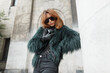 Fashionable beautiful glamorous girl model with cool sunglasses in a fashion shaggy jacket with gloves and a leather handbag posing in the city near a vintage building