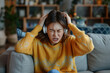 Dissatisfied and angry young Japanese woman sitting on sofa at home and covering ears from excessive noise