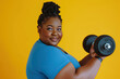 Young happy chubby overweight plus size big fat fit African American woman wear blue top warm up training hold in hand dumbbell look camera isolated on plain yellow background