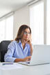 © insta_photos - Vertical photo of busy middle aged business woman in glasses using laptop working in corporate office. Mature professional businesswoman employee at work, female executive manager looking at computer.