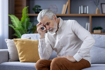 A lonely and upset senior gray-haired man is sitting on the sofa at home, leaning his head thoughtfully on his hand