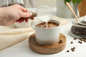 Wall Mural - Woman pouring milk into cup with coffee at white wooden table, closeup