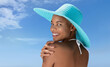 Portrait of a woman wearing a blue sun hat on the hot summer beach, enjoying a holiday by the sea. Seen from behind, with her hand on her shoulder, smiling as she looks into the camera