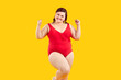 Happy plus size female fashion model in swimwear. Studio shot of cheerful positive young fat woman wearing red one piece swimsuit standing isolated on yellow background, smiling and doing yes gesture