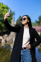 Sticker - Korean woman smiles as she takes a selfie with her cellphone in the park.