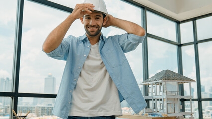 Portrait of smart architect engineer wear safety helmet while sitting on table with house model, project plan and architectural equipment surrounded by skyscraper view. Civil engineering. Tracery