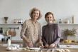 Intergenerational family, two women younger and older age cooking together in modern kitchen at home, standing at dining table, making dough, prepare cookies dessert for holiday, smile, look at camera
