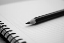A Black Pencil Sits On Top Of A Spiral Notebook. The Notebook Is Open To A Blank Page. The Background Is White.
