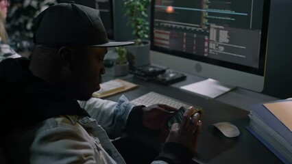 Medium close-up of young African American male mobile application developer in hoodie and baseball cap sitting at desk in office, writing computer code, then checking for errors on smartphone
