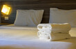 Stack of white towels whick folded neatly on top of bedspread. White bed is neatly made with the covers pulled up tight in hotel room.