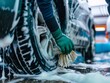 A person is washing a car tire with a green brush. Tire is covered in water and soap. Blue car wash with white soap foam. Auto care service. Car cleaning service concept. Vehicle cleaning service. 