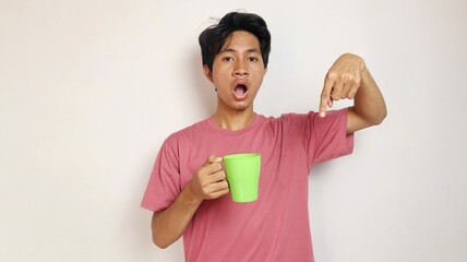 Wall Mural - young shocked asian man posing pointing to the bottom side holding a cup on an isolated white background. wearing a red shirt