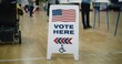 © Framestock - Vote here sign on floor. Woman with spinal muscular atrophy in electric wheelchair comes to vote in polling station. Political races of US presidential candidates. National Election Day. Dolly shot.