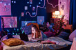 © AnnaStills - Modern teen girl lying on bed in her room decorated with posters and neon lights using laptop while doing homework late in evening