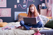 Red-haired Caucasian girl sitting on bed in her bedroom doing homework with use of laptop and internet, copy space
