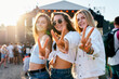 © artiemedvedev - Group of young women at sunny seaside music festival. Smiling friends make peace signs, enjoy live concert. Casual summer fashion, sunglasses, carefree vibe on sandy shore during sunset party.