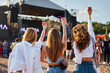 © artiemedvedev - Group of women with colorful drinks at beach festival, arms raised in celebration during summer music event. Friends enjoy sunny day, live performance, seaside weekend party atmosphere.