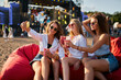 © artiemedvedev - Group of women enjoy music fest on beach, lounging on bean bags. Casual attire, sunglasses, sipping drinks, one takes selfie. Warm daylight highlights relaxed, fun atmosphere of outdoor summer event.