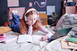 Teen schoolgirl wearing headphones lying on bed in front of laptop making notes during online class, copy space
