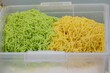Two bowls of noodles, one green and one yellow, sit in a clear container. The green noodles are piled on top of the yellow noodles, creating a colorful and appetizing display